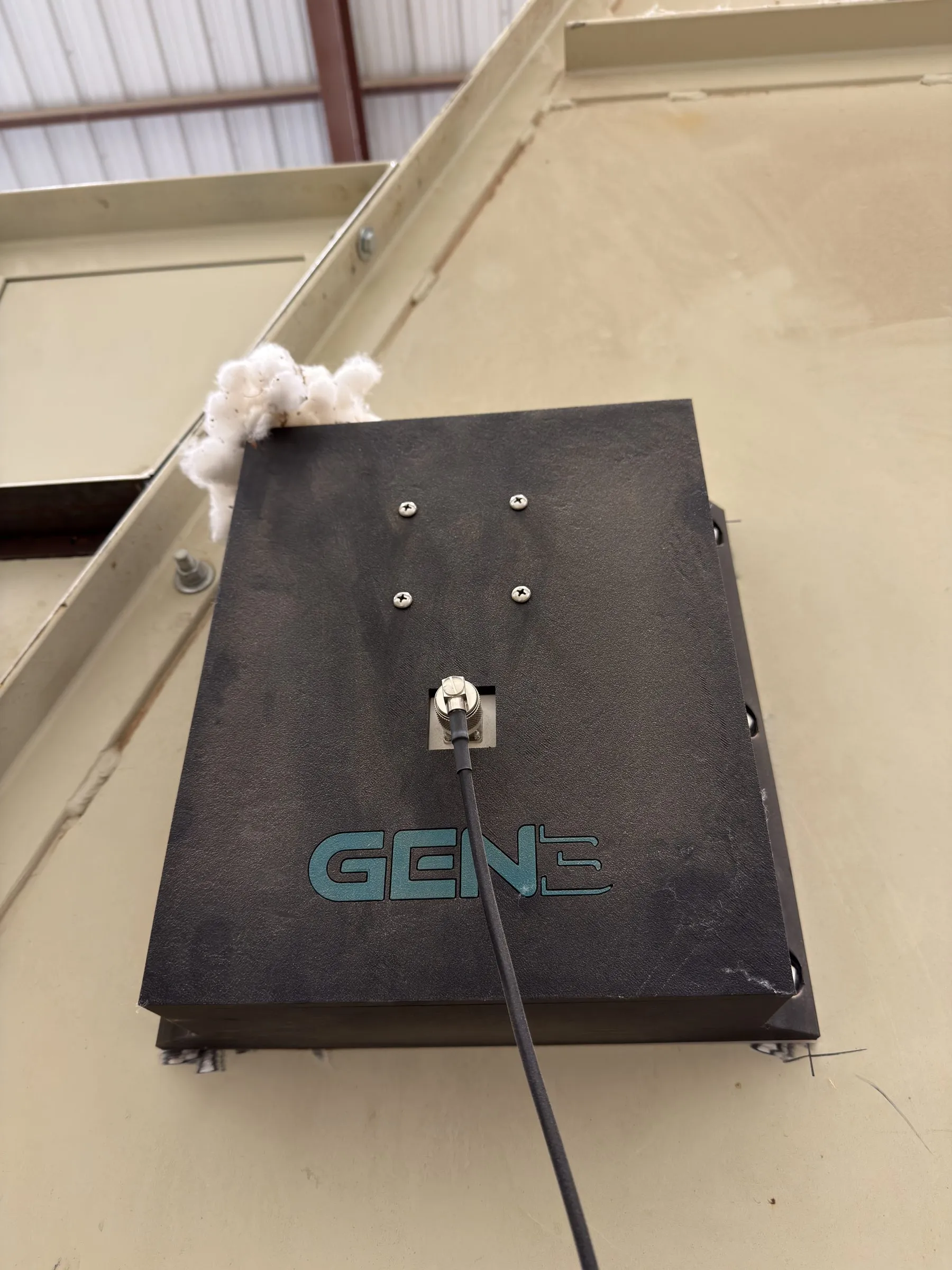A dark Gen 3 Dynamics antenna plate bolted to the wall of a module feeder, a coax lead running from its face, with seed cotton caught on one edge
